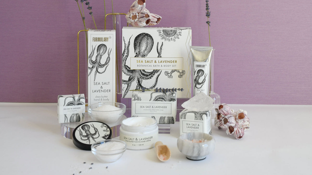 Sea salt and lavender-themed bath products set against a dark backdrop. Includes a boxed set, candle, body scrub, and soap, accented with lavender sprigs and sea salt. Packaging features an octopus illustration.