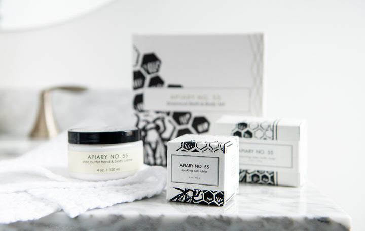 A bathroom countertop displaying Apiary No. 55 products, including a jar of shea butter hand and body cream and two boxes of sparkling bath tabs. The products feature a white and black minimalist design with honeycomb patterns.