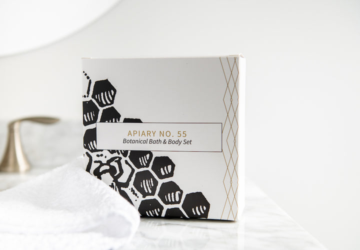 A botanical bath and body set in a box with geometric and floral designs stands on a marble surface next to a white towel and a metallic faucet in the background. The text on the box reads "Apiary No. 55: Botanical Bath & Body Set".